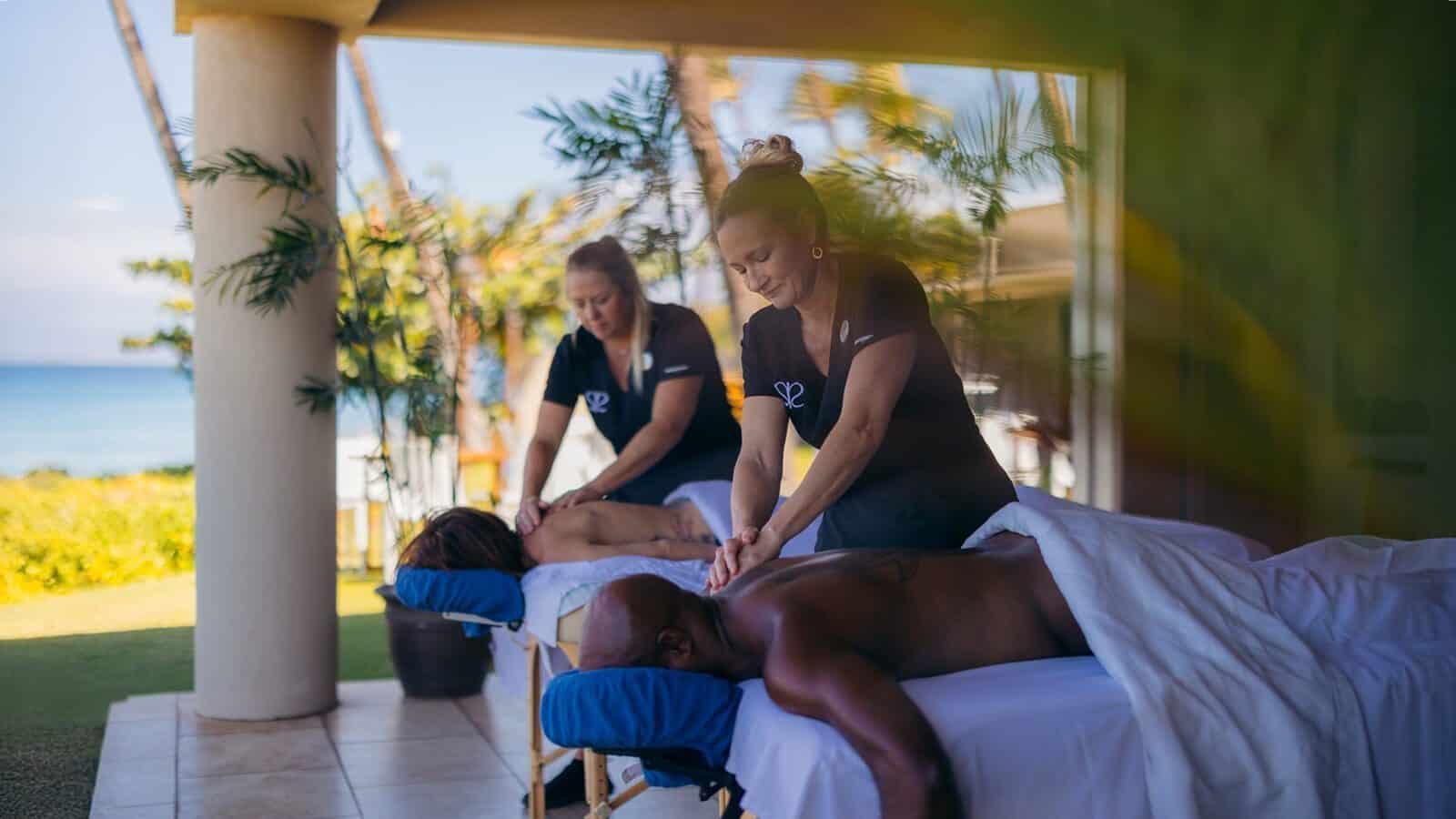 Two massage therapists give outdoor massages to a man and a woman on separate tables, set against Lahaina’s lush, tropical backdrop with breathtaking sea views—a serene escape that celebrates resilience and renewal.