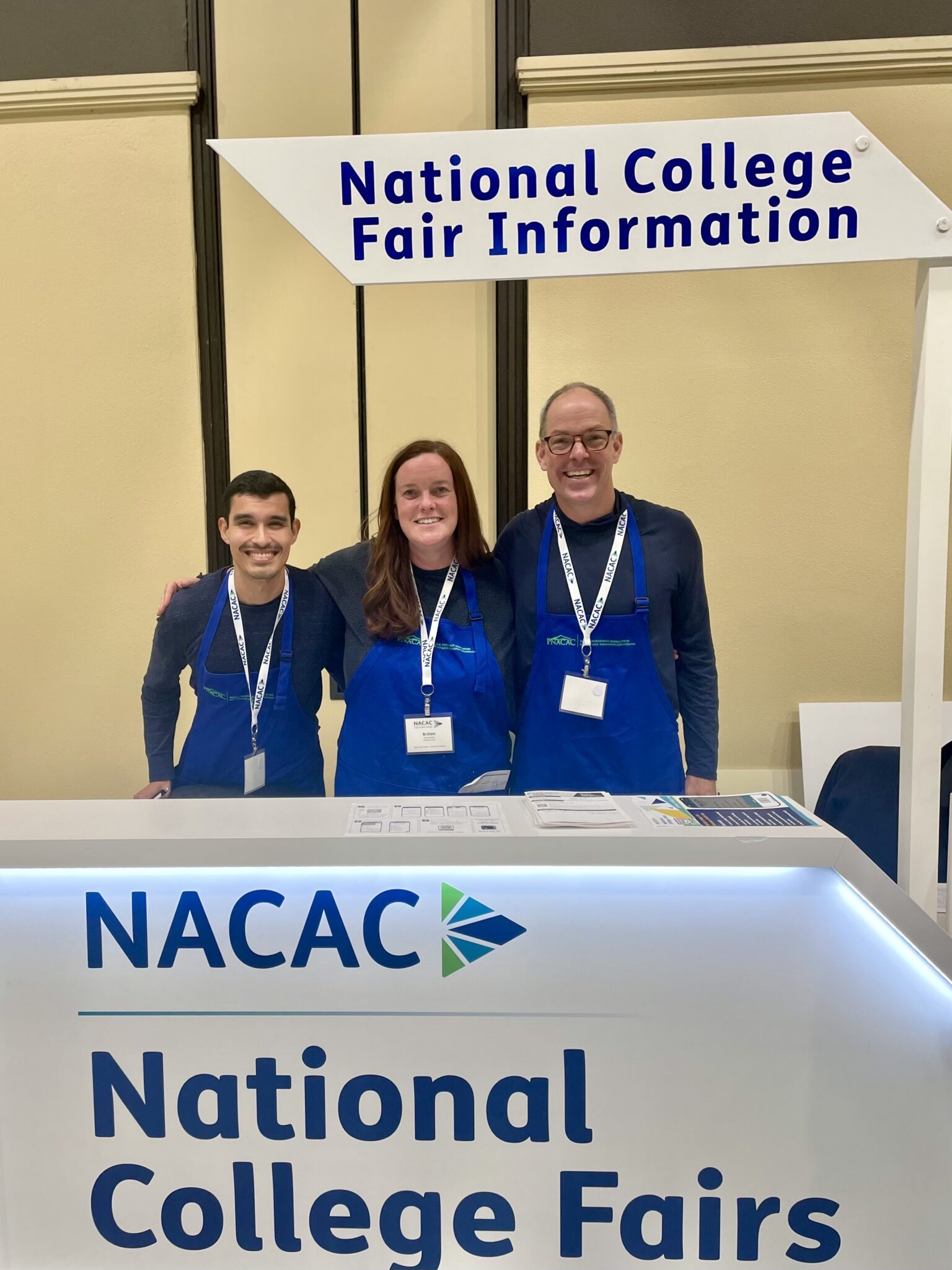 Three people wearing blue aprons and name badges stand behind a NACAC National College Fairs information booth, ready to share insights about UPrep private school and college admission at the event.