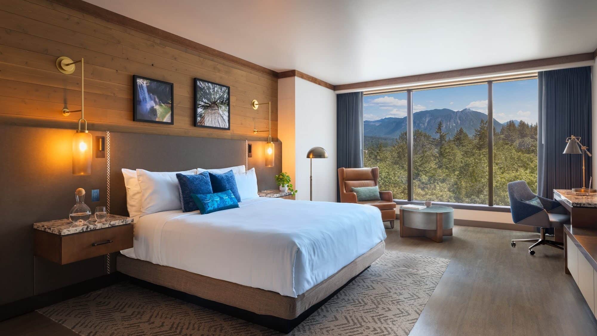 Modern hotel room at Snoqualmie Casino & Hotel with a large bed, armchair, desk, and floor-to-ceiling window overlooking trees and distant mountains—perfect for relaxing after dining or gaming.