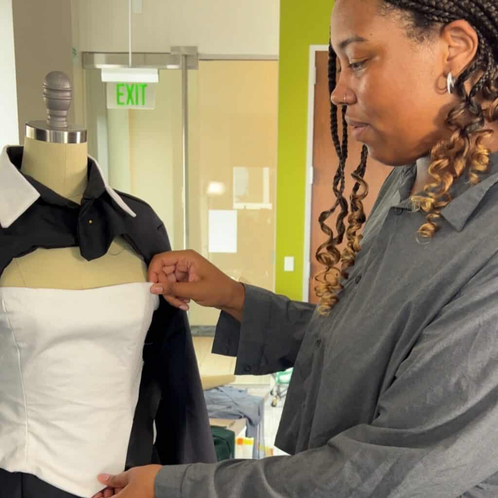 A person adjusts a white fabric piece on a dress form that is dressed in a black collared shirt inside a modern workspace.