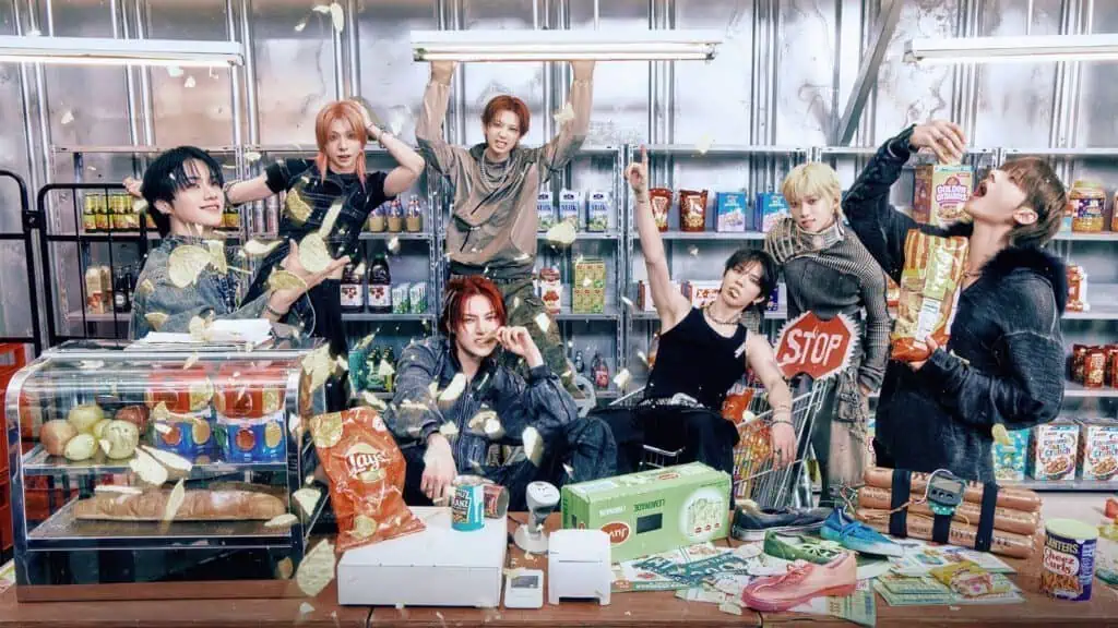 Seven young people pose in a snack-filled convenience store, with chips flying in the air and various food items scattered on the counter and shelves behind them.