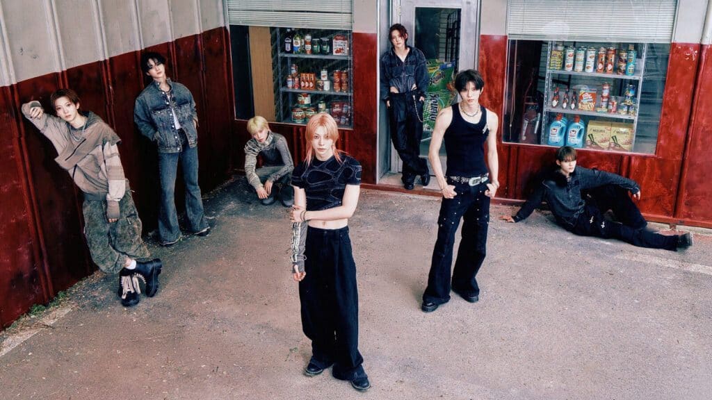 Six people stand and sit in a group outside a building with large windows displaying various products, posing casually in front of a red and gray wall.