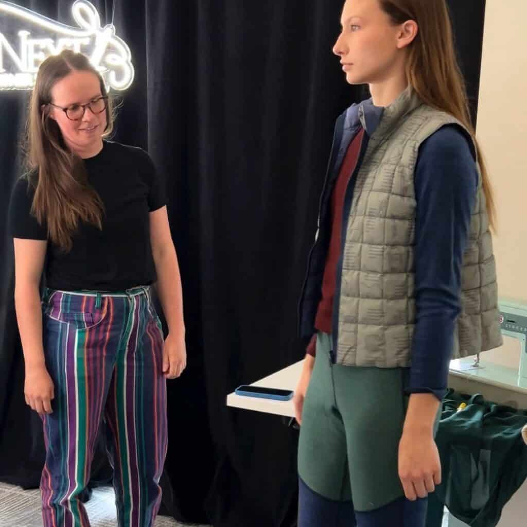 Two women stand indoors; one wears a black shirt and striped pants, while the other wears layered athletic clothing and a quilted vest. A sewing machine is partially visible on the right.