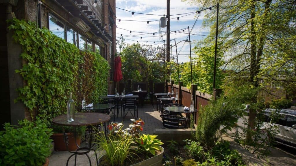 Outdoor restaurant patio with black metal tables and chairs, string lights overhead, and greenery lining the walls and surrounding area on a sunny day.