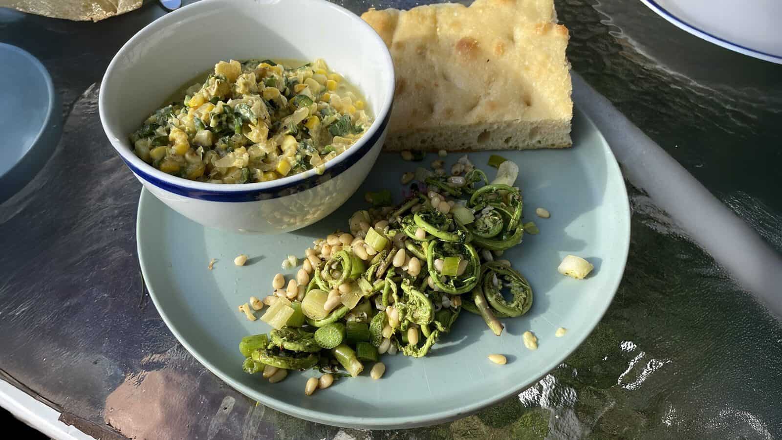 A plate with sautéed fiddlehead ferns and pine nuts, a piece of focaccia bread, and a bowl of vegetable mixture create a fresh Trail to Table experience, all beautifully arranged on a glass table.