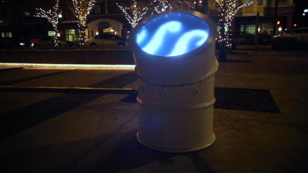 A white metal barrel with a glowing blue screen on top, displaying a wavy pattern, stands on a sidewalk at night during an art walk in Seattle, with lit trees illuminating the background.