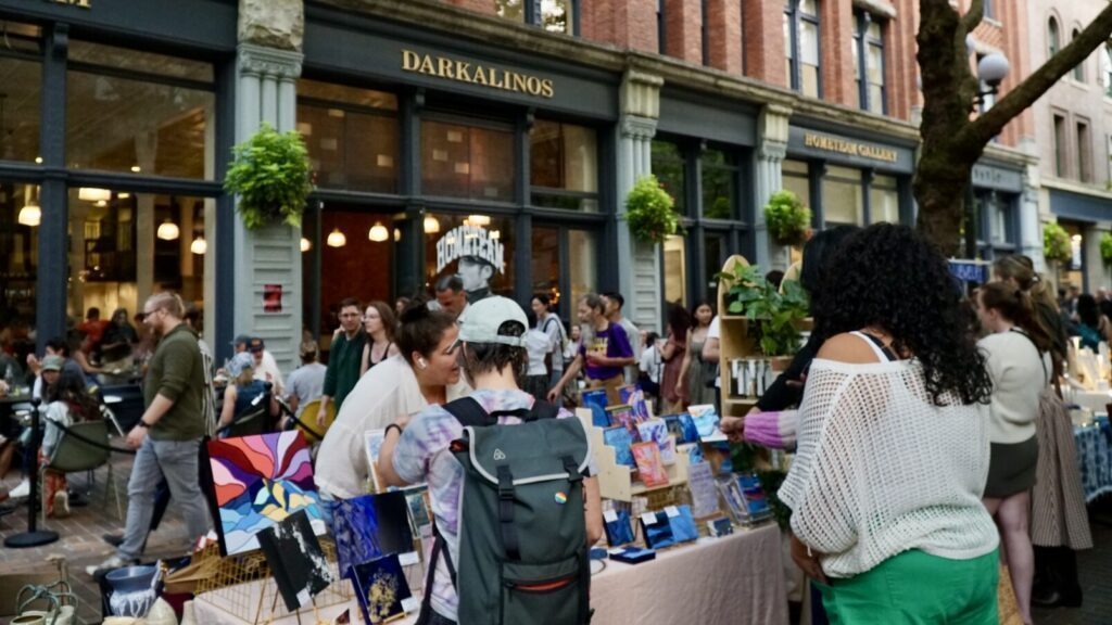 People browse artwork and crafts displayed on tables at an outdoor market in front of brick buildings and a restaurant named “Darkalinos.”.