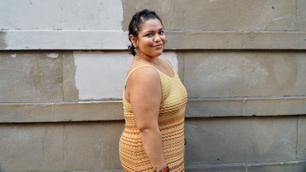 A woman in a yellow and orange sleeveless dress stands in front of a beige stone wall, looking at the camera with a slight smile.
