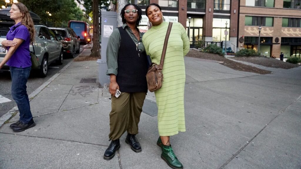 Two women stand side by side on a city sidewalk, one in green overalls and black top, the other in a light green dress, both wearing boots and looking at the camera.