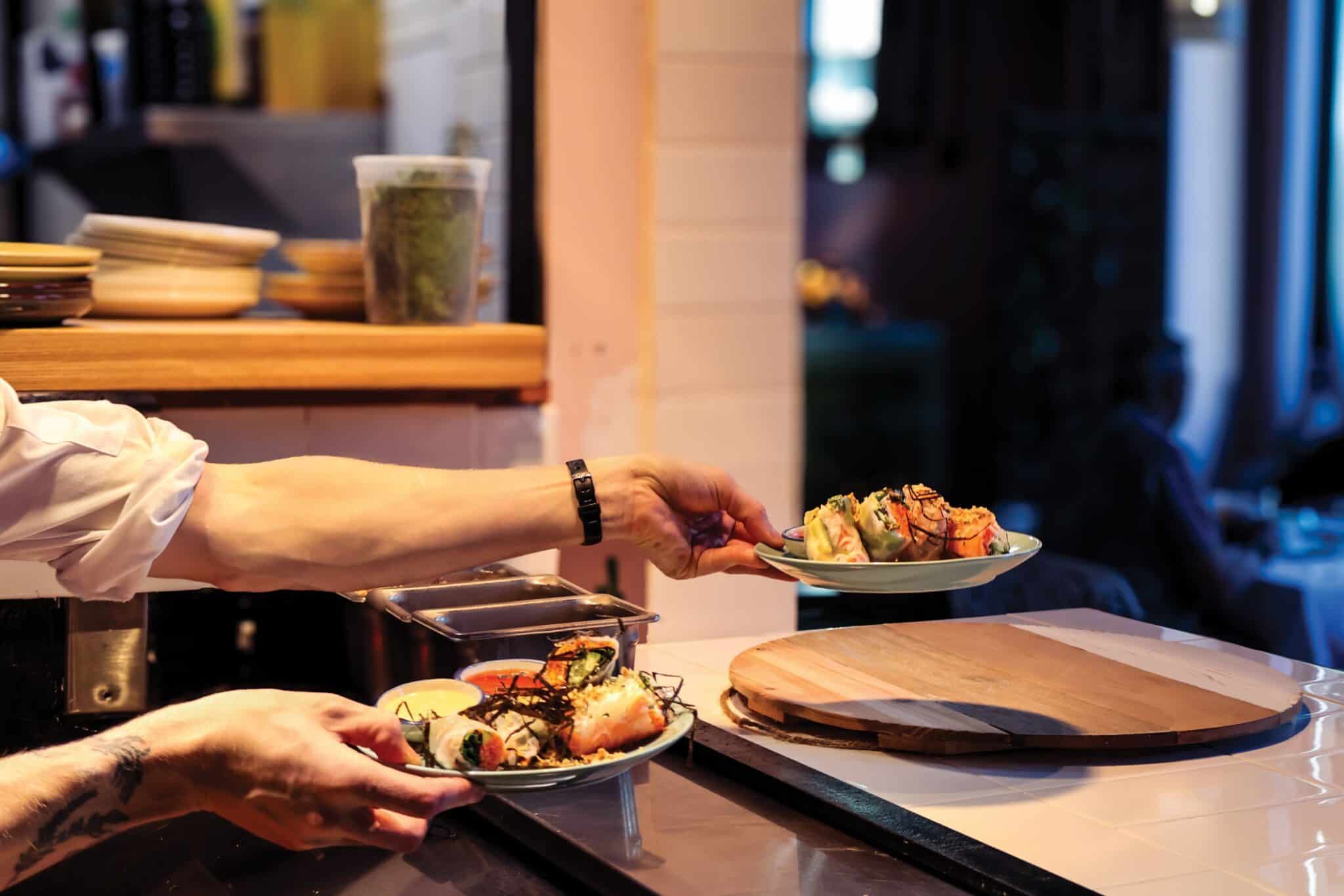 A person places two plates of prepared food on a counter in La De Soil’s restaurant kitchen, with more dishes and ingredients visible in the background—a great spot for those seeking vegetarian restaurants or wondering where to eat in Seattle.