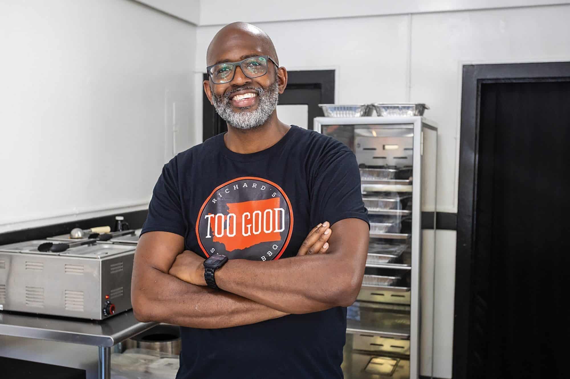 Man with glasses and a beard, wearing a "Too Good" t-shirt, stands with arms crossed in a commercial kitchen—perhaps he's the chef behind La De Soil, one of the top vegetarian restaurants on any "Where to eat in Seattle" list.
