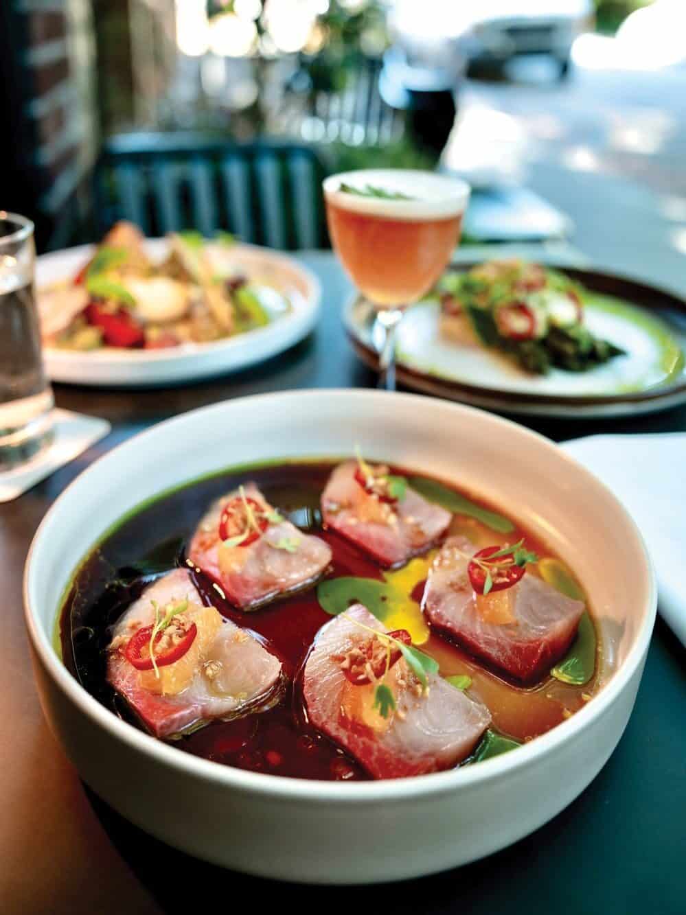 A bowl of sashimi-style fish slices in sauce, garnished with herbs and chili, sits on a table—a true masterclass in presentation—surrounded by additional plated dishes and a cocktail in the background.