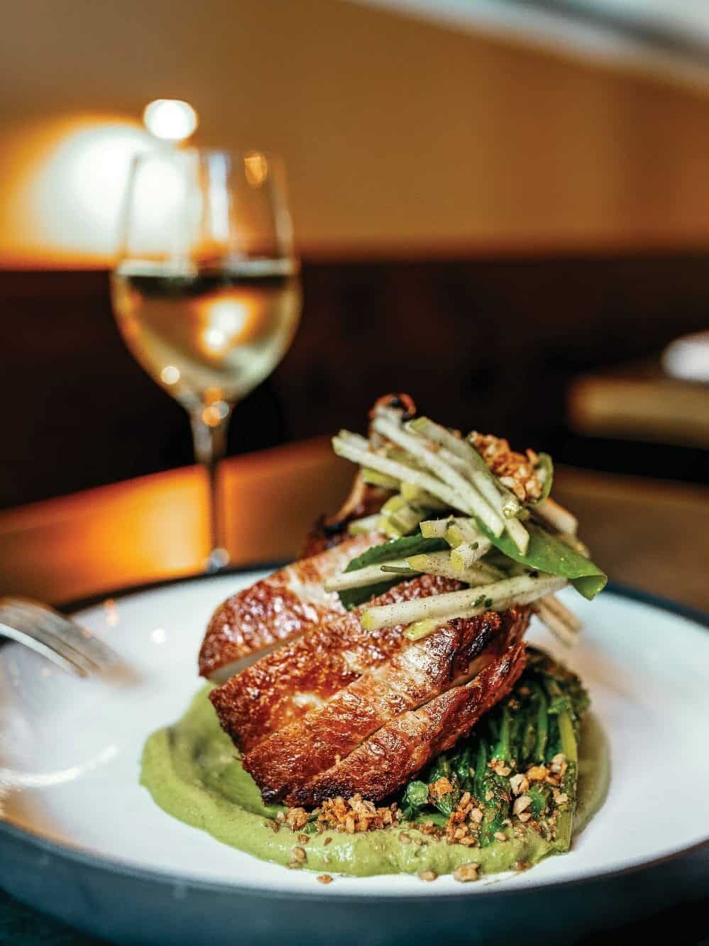 A plated dish featuring seared meat atop green puree and vegetables, garnished with thinly sliced apples—a true masterclass in presentation—with a glass of white wine in the background.