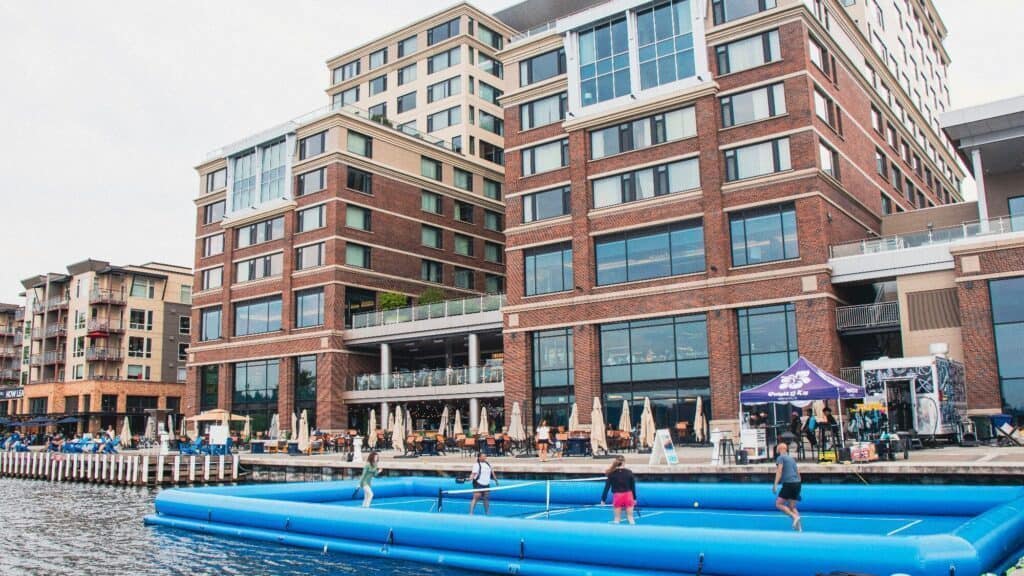 Several people play tennis on a floating court in a waterfront area with a large brick hotel and outdoor dining in the background.