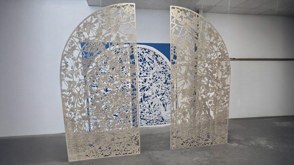 Two intricately cut metal panels with botanical designs stand slightly apart in front of a blue and white artwork on a wall in a minimalist gallery space.