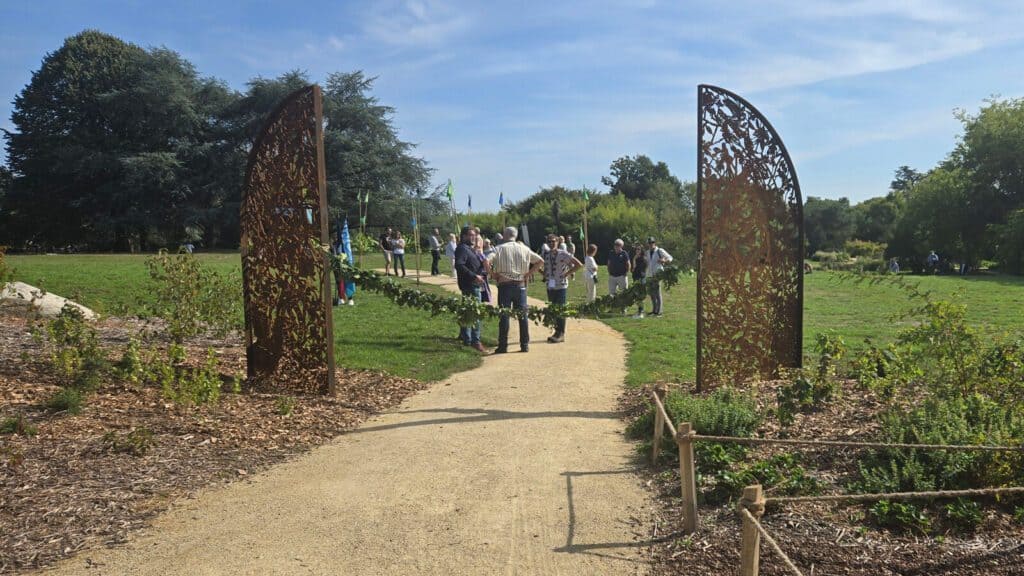 A group of people gather near a decorative gate adorned with greenery on a dirt path in a park under a clear blue sky.