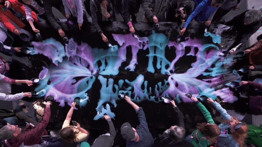During an art walk in Seattle, people stand around a large black table, creating symmetrical patterns with blue and purple powders—colors that celebrate the vibrant LGBTQIA+ community—as they spread them outward with spoons.