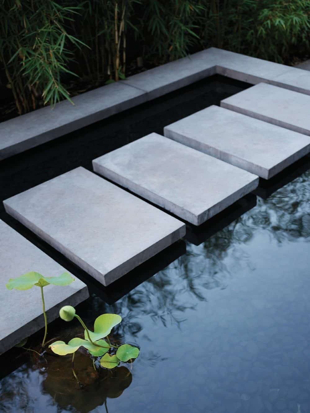 Rectangular stone stepping stones form a path across a shallow water feature, with green lotus plants and bamboo in the background—a serene touch perfect for custom-built homes.