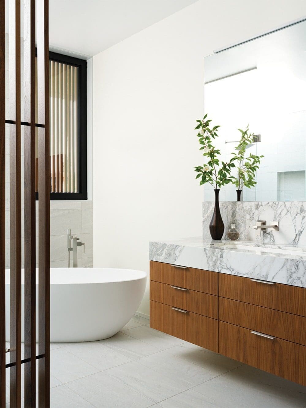 Modern bathroom with a freestanding white bathtub, marble countertop, wooden vanity drawers, large mirror, and tall vases—perfect for increasing home remodel value in custom-built homes.