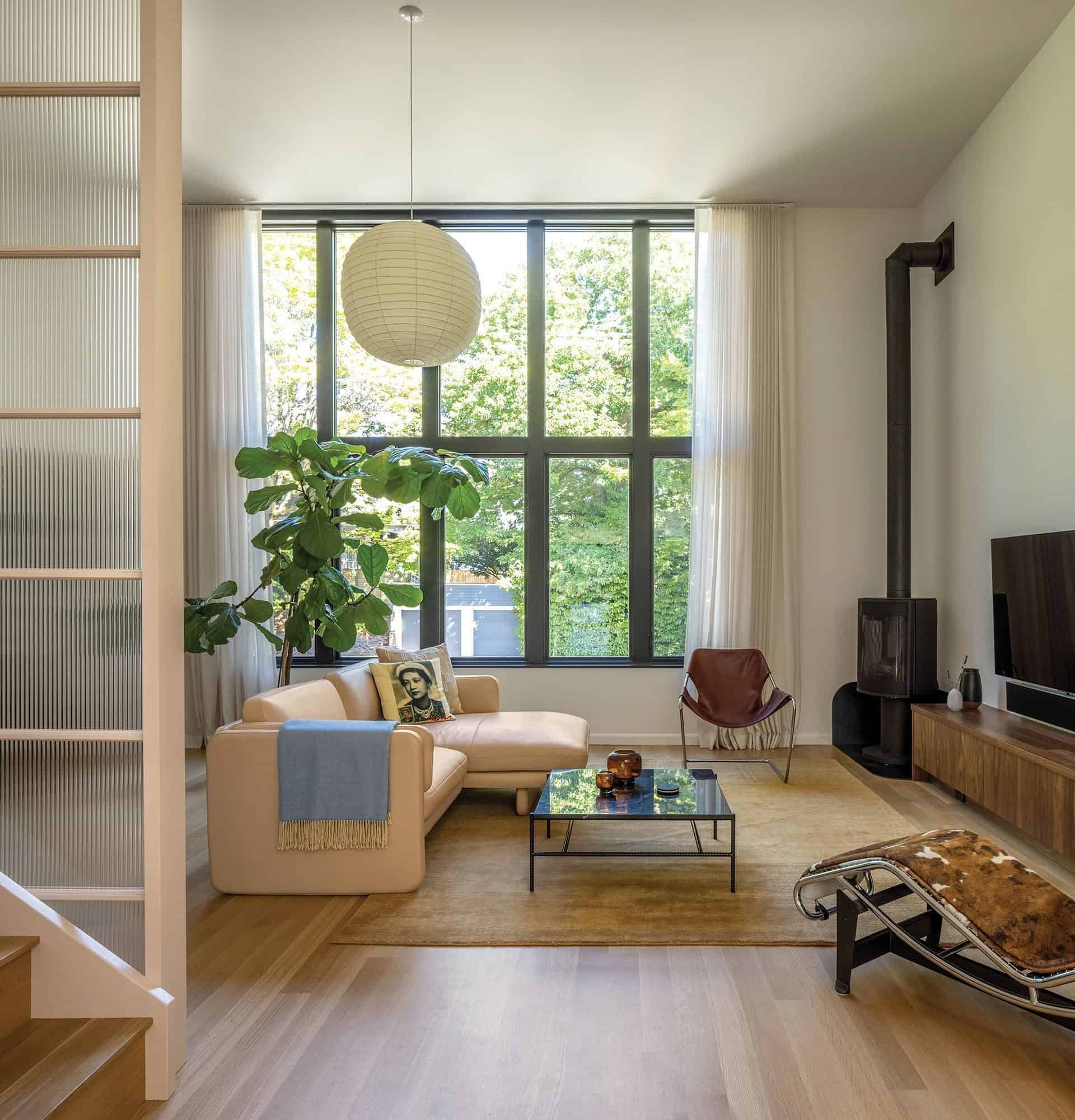 Modern living room with large windows, a tan sectional sofa, indoor plant, TV on a wooden console, and glass coffee table. Neutral tones and natural light fill the space—perfect for building community in a welcoming setting.