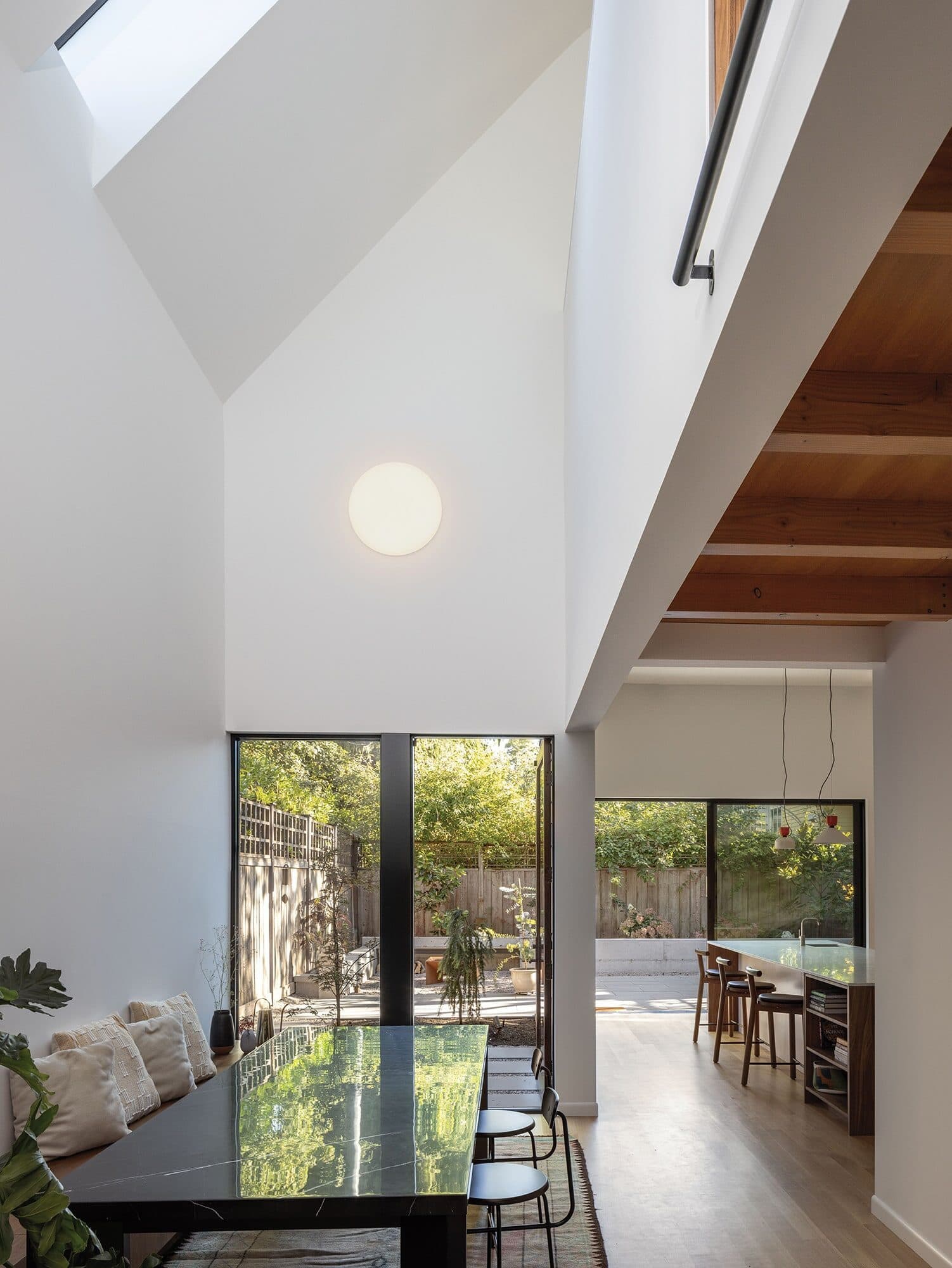 Modern dining area with high ceilings, skylights, a marble table, built-in bench seating, and large windows overlooking a garden; the design reflects community blueprints, with an open kitchen area visible for building community connections.