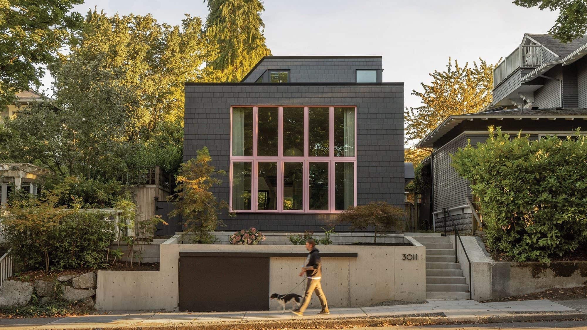 A modern house with large pink-trimmed windows, gray exterior, and concrete entry sits behind landscaping. A person walks a dog on the sidewalk, reflecting the spirit of building community in this inviting neighborhood.