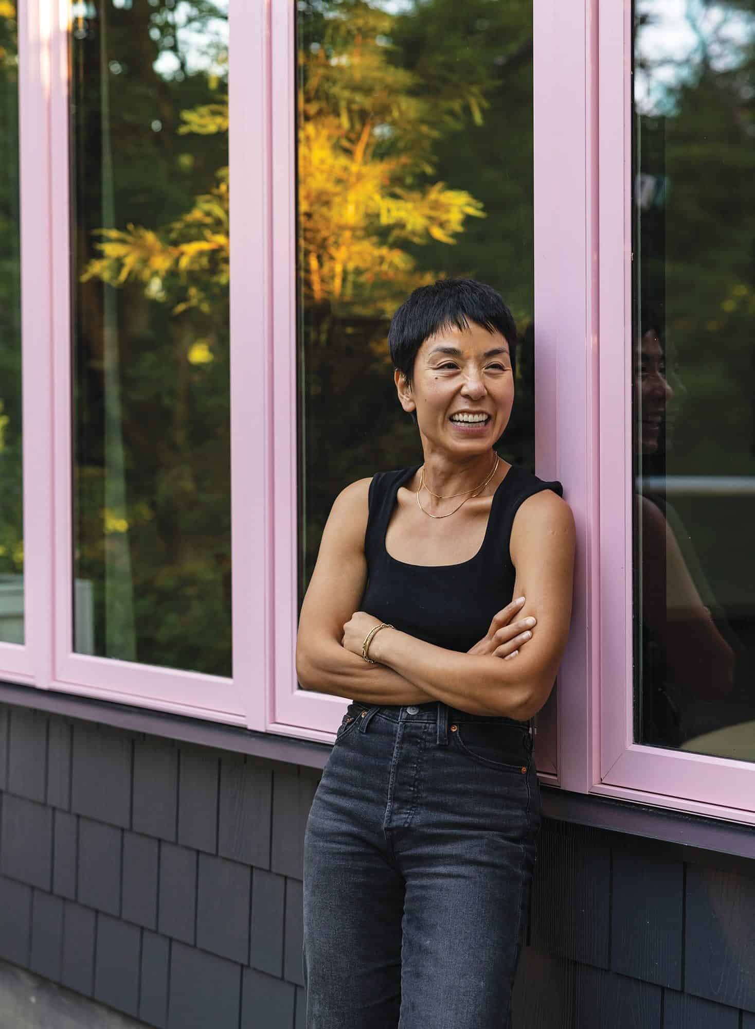 A person with short hair wearing a black tank top and dark jeans stands smiling with arms crossed in front of large pink-framed windows, embodying the spirit of building community.