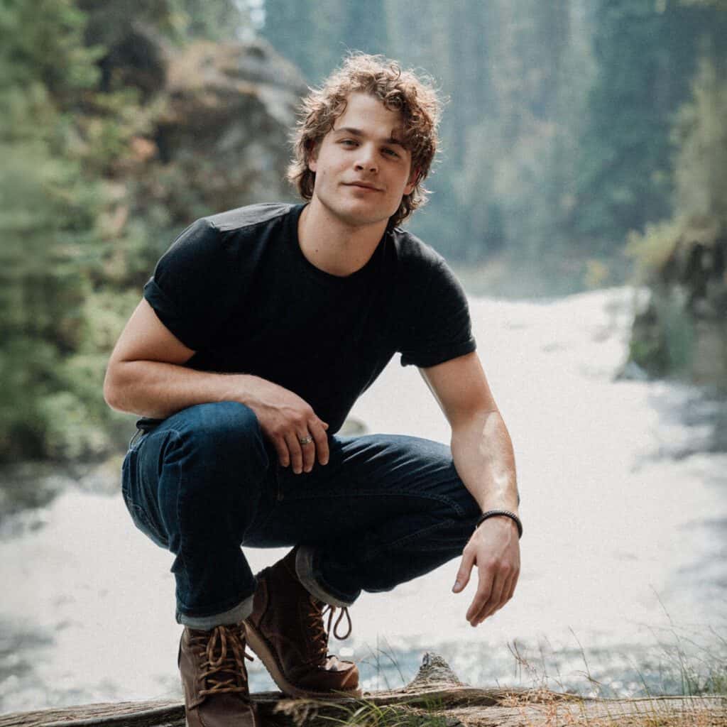 A young man in a black t-shirt and jeans crouches on a log beside a river in a forested area.