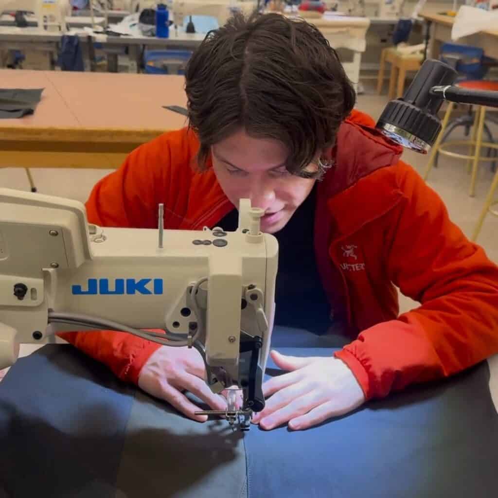 A person in a red jacket uses a Juki industrial sewing machine to stitch black fabric in a workshop, reflecting the creativity of young fashion designers preparing pieces for the runway.