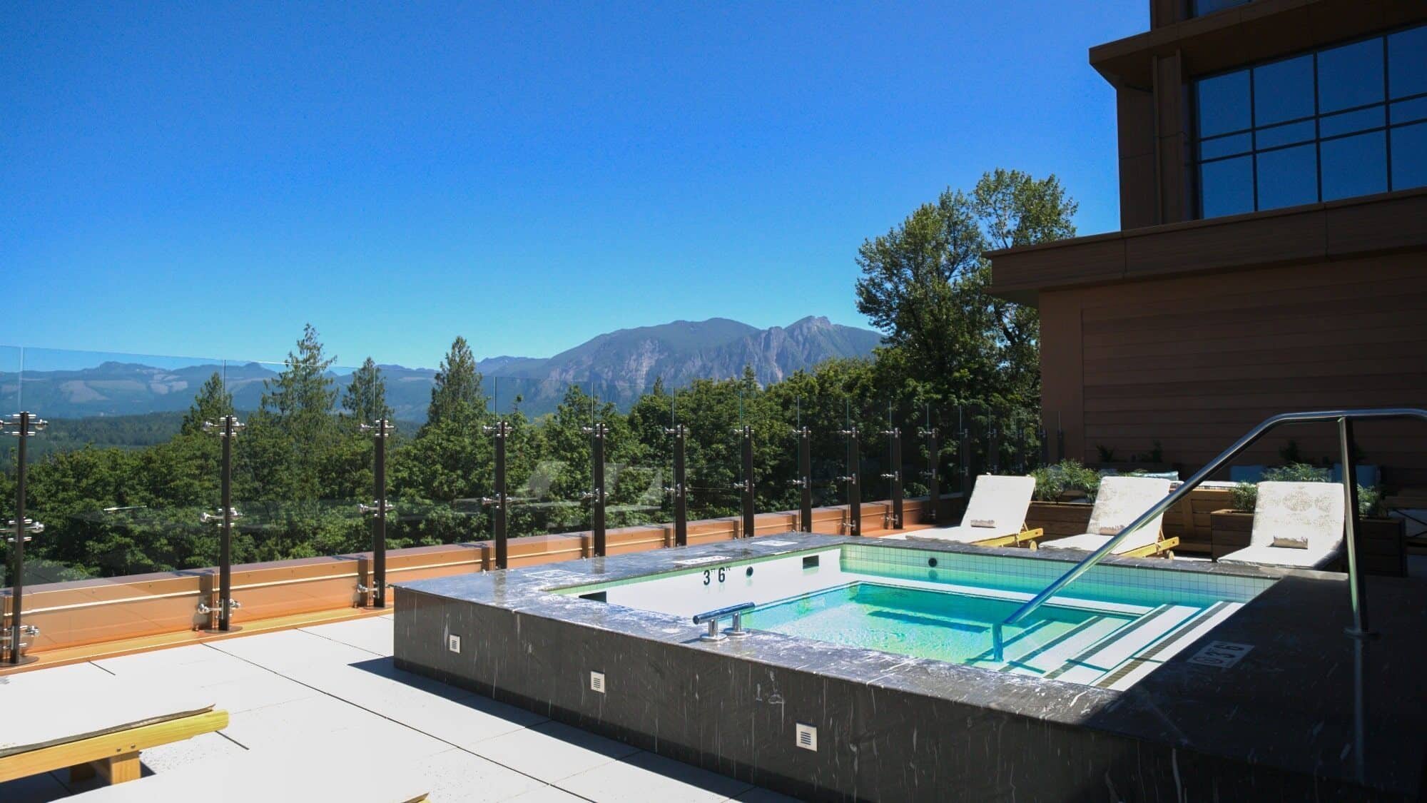 A modern outdoor hot tub with lounge chairs on a rooftop patio at Snoqualmie Casino & Hotel, surrounded by glass panels and overlooking a forested mountain landscape under a clear blue sky. Enjoy stunning views after dining or playing at the casino.
