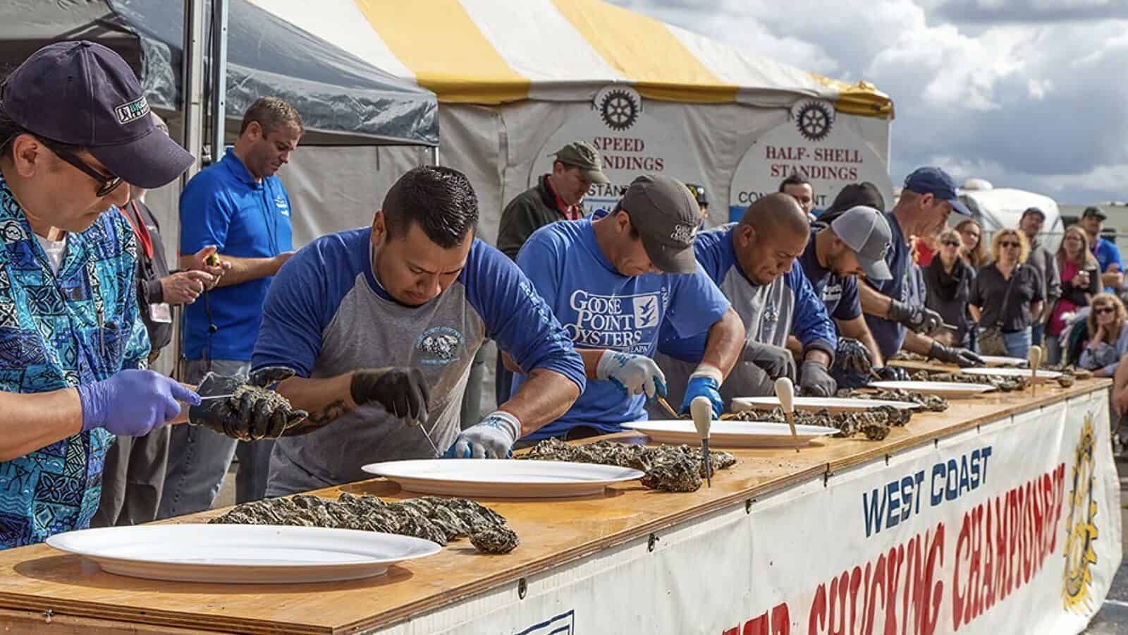 Five men compete in an oyster shucking contest at an outdoor event, each opening oysters and placing them on plates, showcasing classic Fair Fare style while spectators watch in the background.