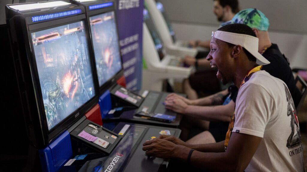 A man enthusiastically plays an arcade fighting game on a gaming machine, with other players seated beside him, at a gaming event.