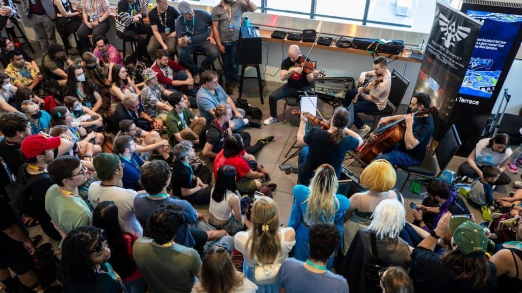 A string quartet performs for a large seated audience at an indoor event, with people attentively listening and some wearing cosplay costumes.
