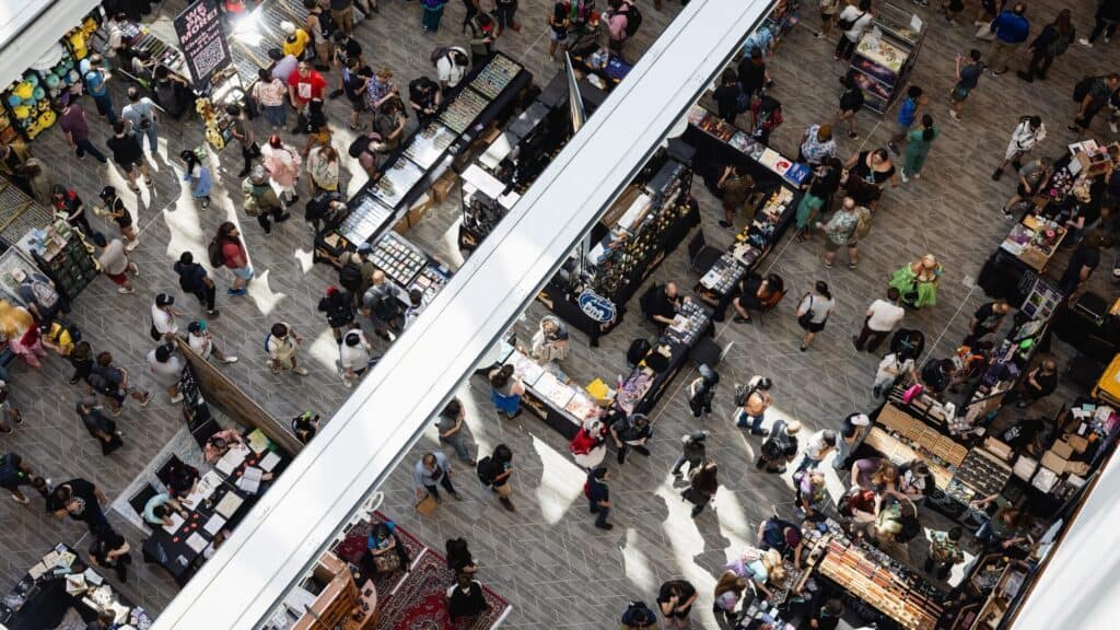 Crowds of people walk among booths and tables at an indoor convention or market, viewed from above through white beams.