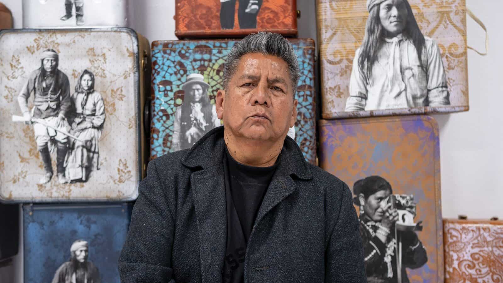 A man stands in front of a wall of decorated suitcases, each featuring historical photos of Indigenous people from Pueblo tribes, reflecting the rich heritage found in the heart of Santa Fe, the City Different.