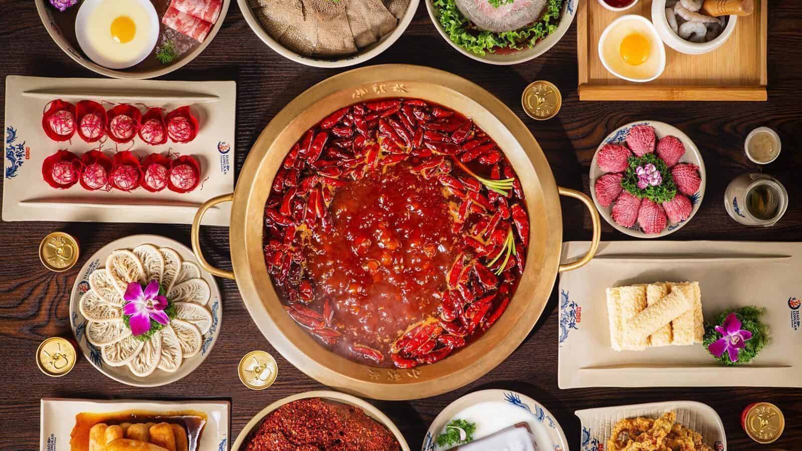 A large pot of spicy hotpot with vegetables, chili peppers, and local tofu is surrounded by various raw meats, seafood, and dipping sauces on a table.