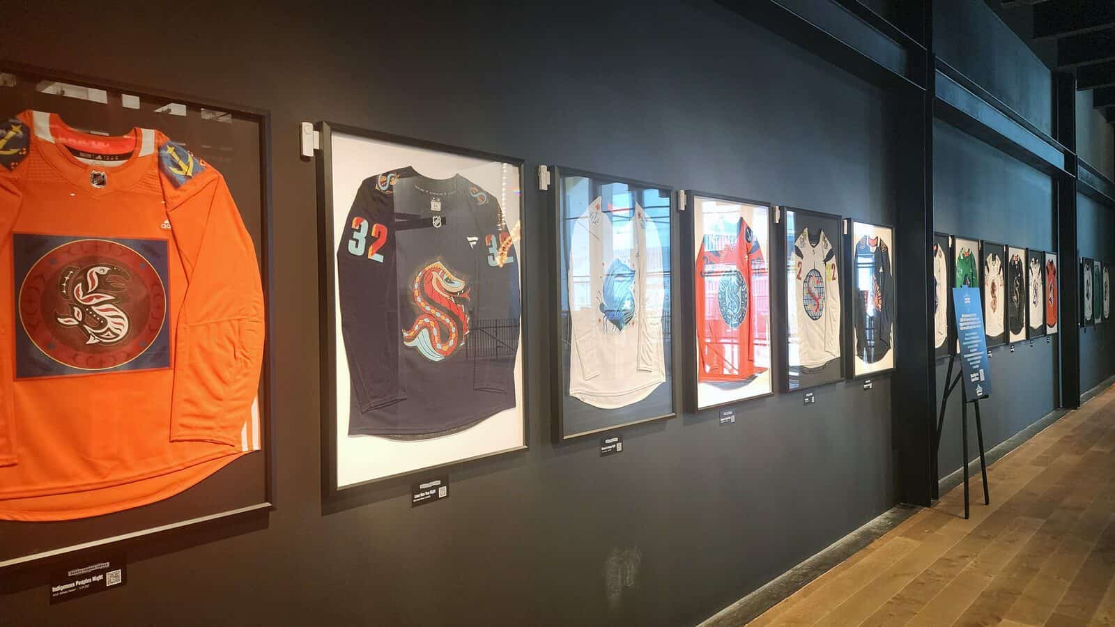 A row of framed artist-designed jerseys, including some from the Kraken, is displayed on a black wall in a museum or gallery setting with wooden flooring.