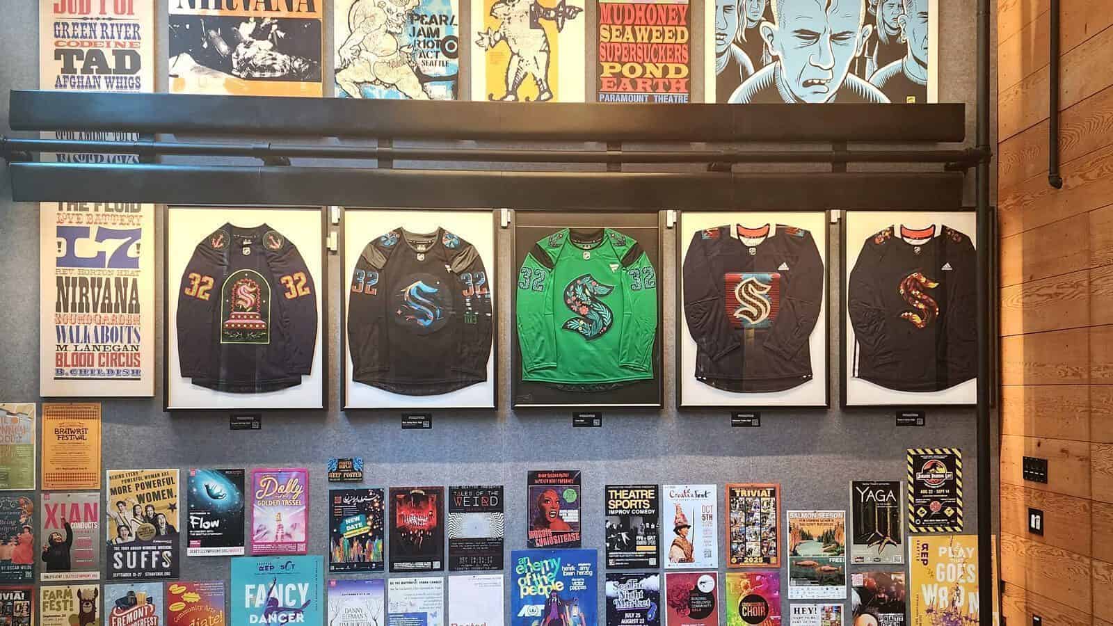 Five framed Kraken artist-designed jerseys are displayed on a wall above a collection of colorful event posters and concert flyers, capturing the vibrant spirit of this season.