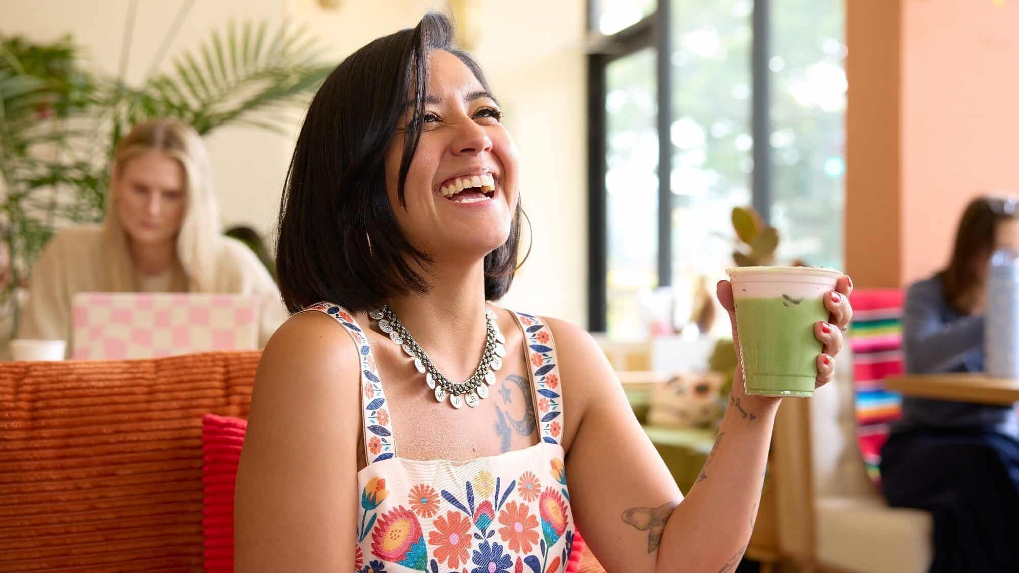 A woman in a floral dress smiles while holding a green smoothie in a brightly lit café, with other people working in the background.
