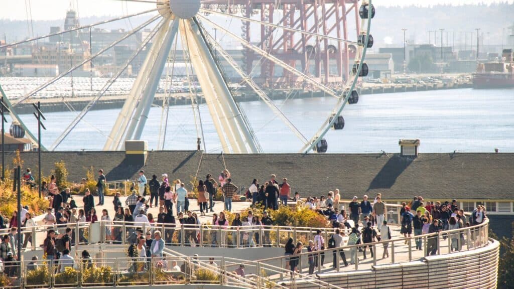 Crowds of people walk along a waterfront promenade with a large Ferris wheel and industrial structures visible in the background on a sunny day.