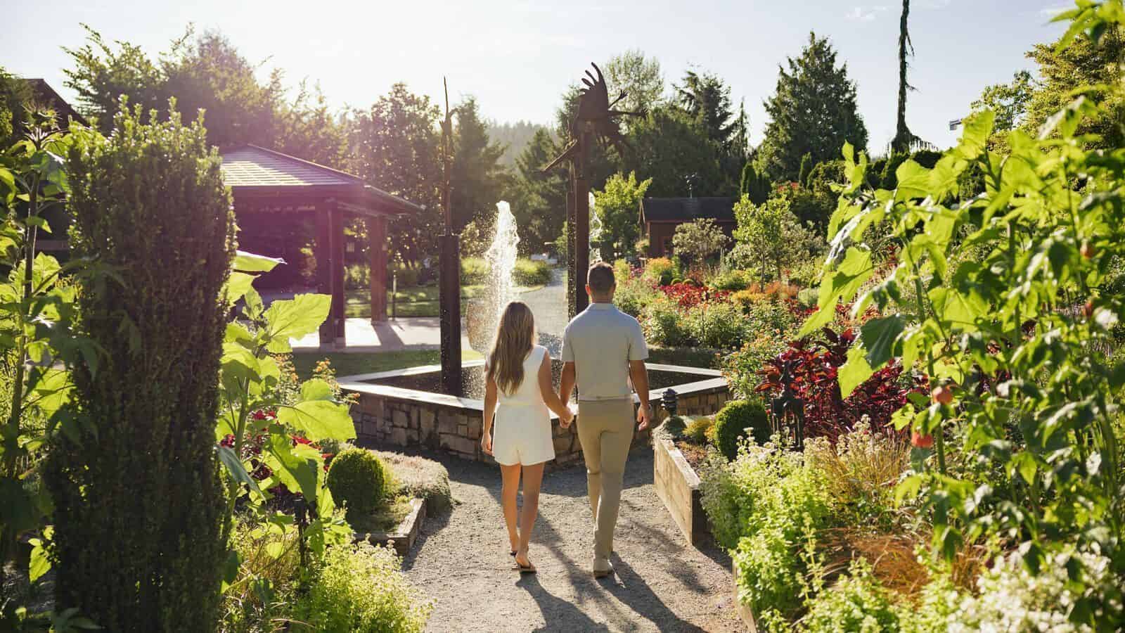 A couple walks hand in hand along a garden path toward a fountain surrounded by greenery and flowers on a sunny day, capturing the relaxation and charm of Woodinville.