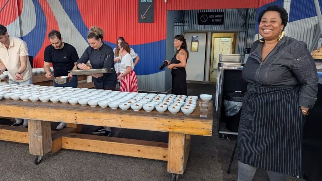 Several people prepare small bowls of food on a long wooden table outdoors, while a woman in an apron smiles at the camera in the foreground.