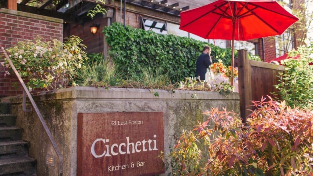 A stone wall sign reads “Cicchetti Kitchen & Bar, 121 East Boston” outside a leafy patio with a red umbrella and a person arranging flowers.