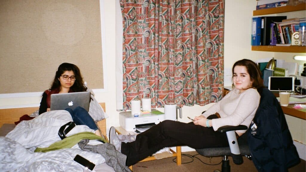 Two young women in a dorm room; one is sitting on a bed using a laptop, while the other sits at a desk with her feet up, looking at the camera.