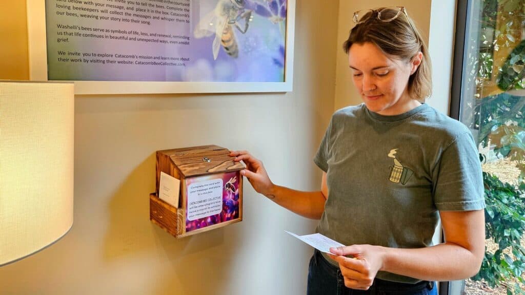 A person stands indoors next to a wooden suggestion box, holding a paper slip and reading it. Informational posters and a window are visible in the background.