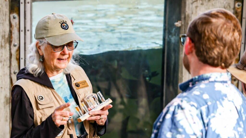 An older woman in a vest and cap holds a small scientific device while talking to a man in a blue patterned shirt in front of a large aquarium window.
