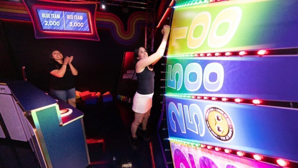 Two people play an arcade-style game, with one person pressing buttons on a vertical score board while the other claps. Digital team scores are displayed in the background.