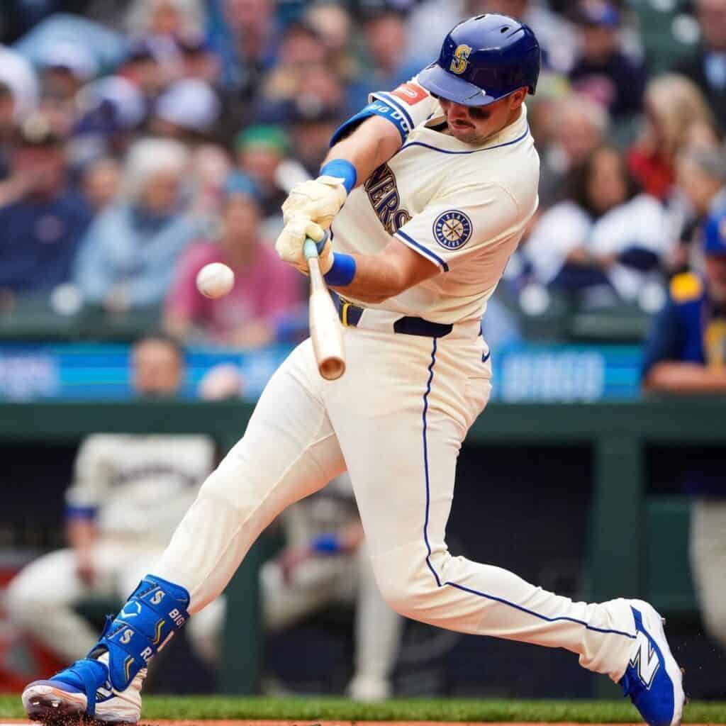 Mariners baseball player in a white uniform swings a bat and makes contact with the ball during a playoffs game, with spectators visible in the background.