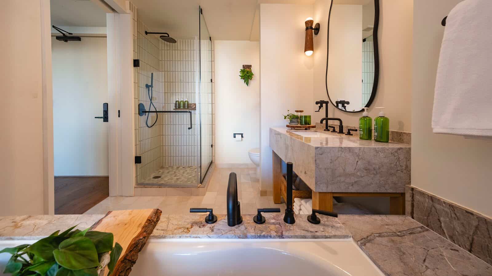 Modern bathroom at 1 Hotel South Lake Union features a glass-enclosed shower, marble countertops, wall-mounted faucets, and green accents. A towel hangs beside the bathtub in this hidden gem of relaxing urban luxury.