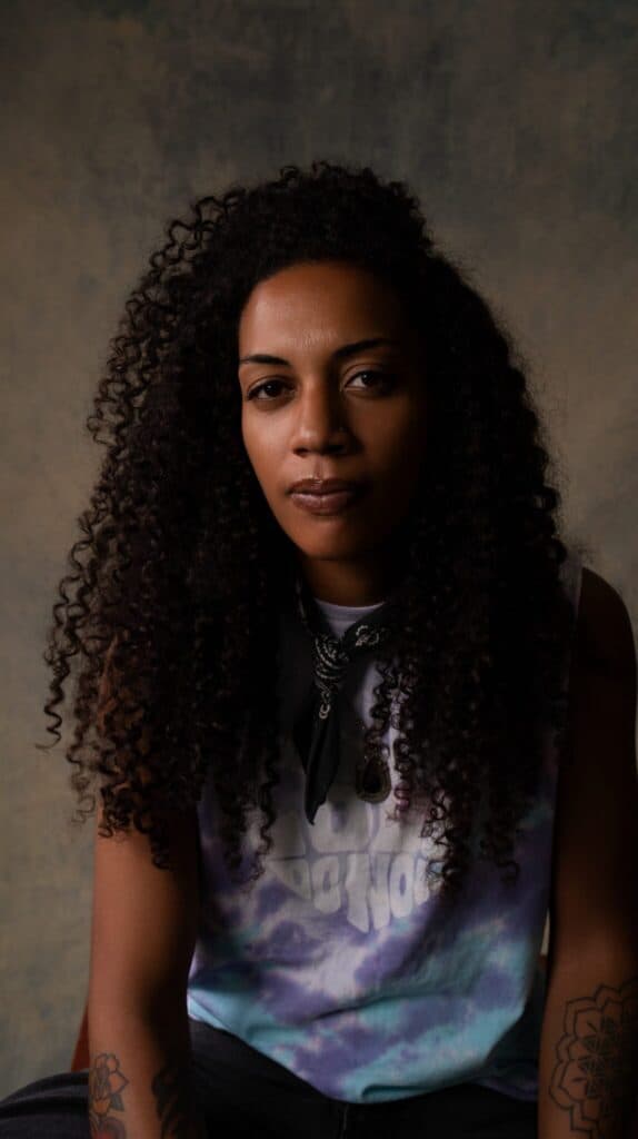 A person with long curly hair, wearing a tie-dye shirt and bandana, sits facing the camera against a neutral background.